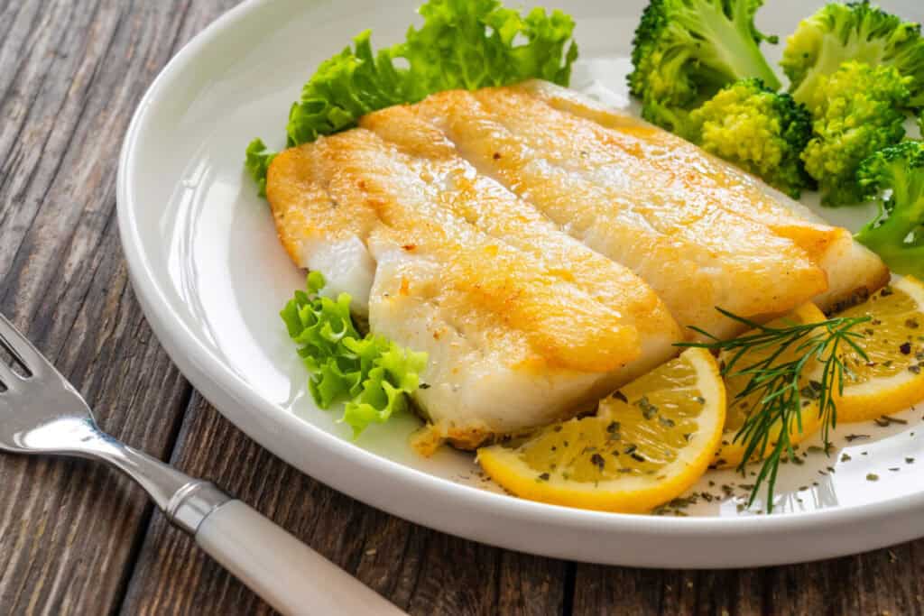 seared halibut fillet with lemon Seared halibut fillet served with lemon slices, fresh lettuce, and steamed broccoli on a white plate over a rustic wooden table
