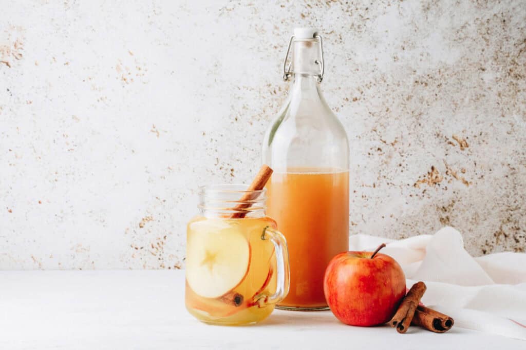 cinnamon honey apple cider vinegar drink Refreshing apple cider vinegar drink with honey, sliced apples, and cinnamon sticks in a mason jar, alongside a bottle of the beverage, a red apple, and cinnamon sticks on a light background.