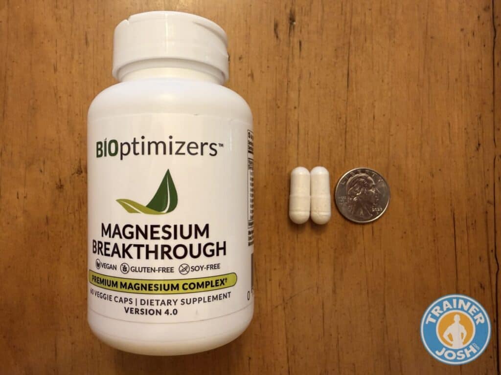 bioptimizers magnesium breakthrough0002 A white bottle of BiOptimizers Magnesium Breakthrough sits on a wooden surface. The label reads “BiOptimizers Magnesium Breakthrough — Premium Magnesium Complex — 60 Veggie Caps — Dietary Supplement — Version 4.0,” with icons indicating vegan, gluten-free, and soy-free. To the right of the bottle are two white capsules placed beside a U.S. quarter coin for size comparison. A blue-and-orange “Trainer Josh” watermark is visible in the lower right corner of the image.*