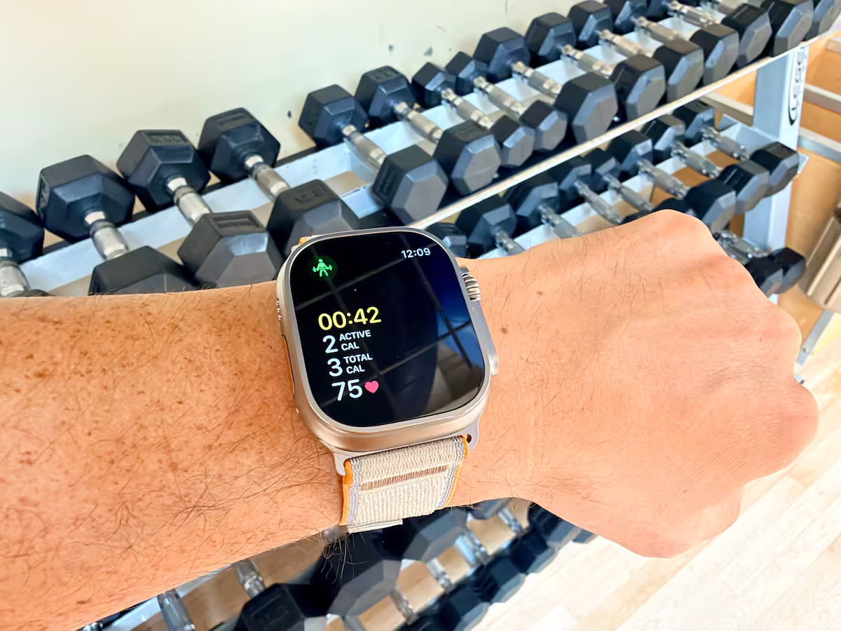 apple watch ultra 4 review wrist gym Apple Watch Ultra 4 on Josh’s wrist displaying strength-training metrics, with a rack of dumbbells in the gym behind it.